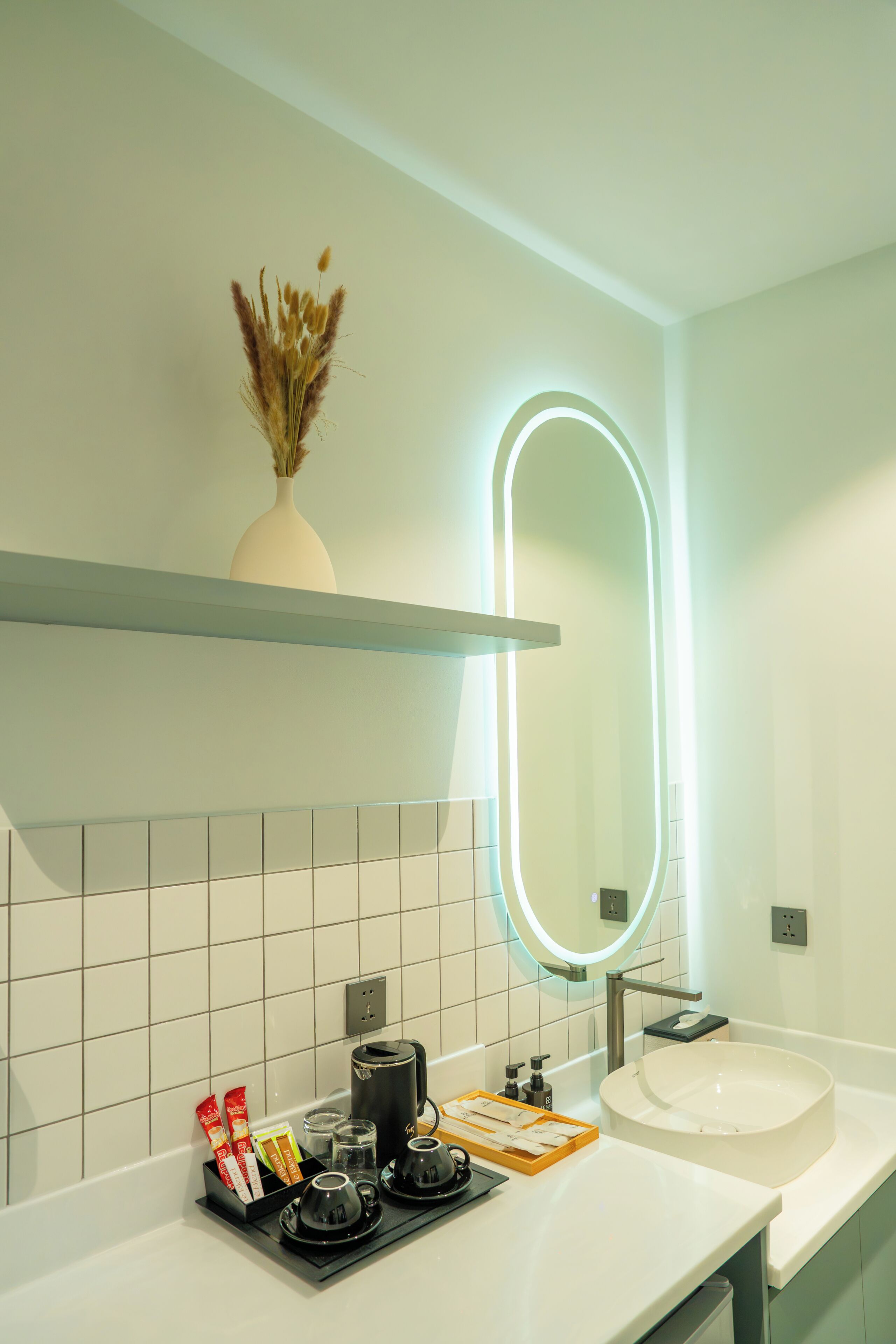 A modern bathroom with a white sink, a mirror with an illuminated frame, a shelf with a vase of dried flowers, and a coffee station with cups and a kettle.