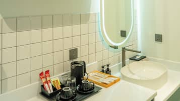 A modern bathroom with a white sink, a mirror with an illuminated frame, a shelf with a vase of dried flowers, and a coffee station with cups and a kettle.