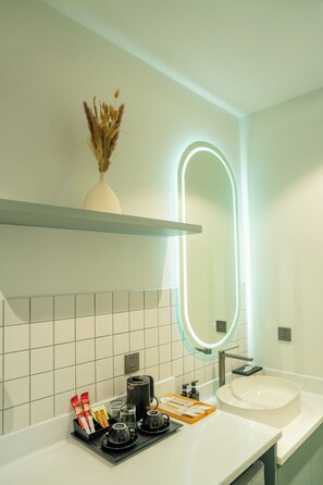 A modern bathroom with a white sink, a mirror with an illuminated frame, a shelf with a vase of dried flowers, and a coffee station with cups and a kettle. - Lé Blend Residence (Krong Poi Pet)