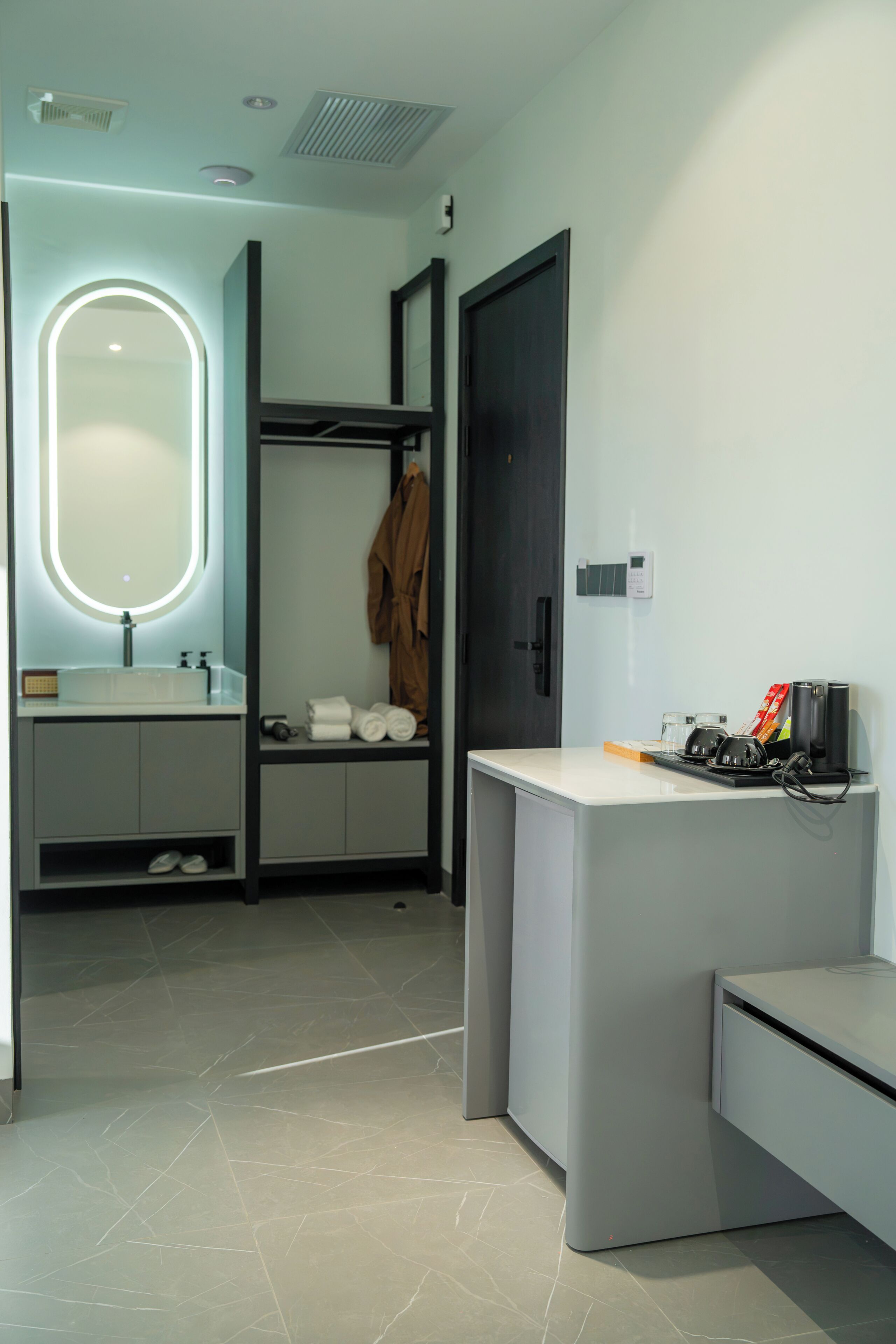 Een moderne badkamer met een wastafel, spiegel, badjas en een klein bureau met een koffiezetapparaat.