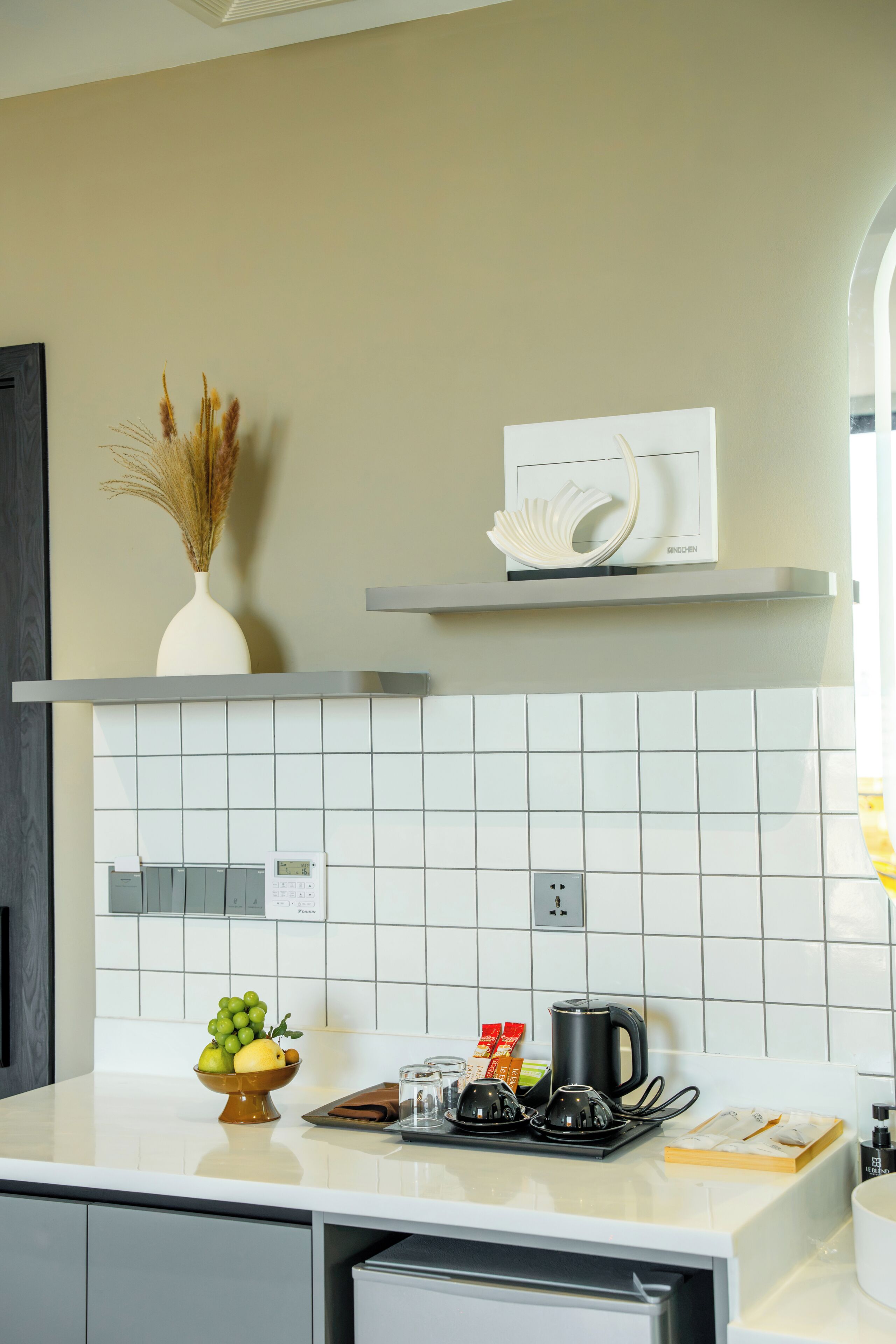 Una cocina moderna con azulejos blancos, encimera blanca, un jarrón blanco con plantas secas, una estantería blanca con un objeto blanco, y un frutero.