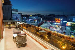 A balcony with wicker furniture, a view of a city street at dusk, and illuminated signs. - Lé Blend Residence (Krong Poi Pet)