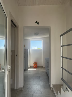 Ein Flur mit Fliesenboden, einem kleinen, orangefarbenen Mülleimer, einem Fenster mit Blick und einem Metallgeländer.