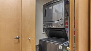 Una safareig compacte amb rentadora i assecadora, una porta de fusta, i una petita prestatgeria amb articles.