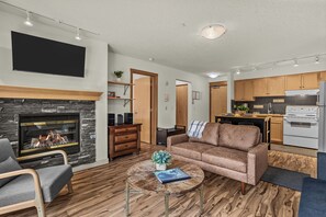 A modern living room with a fireplace, a sofa, a coffee table, and a kitchen area with wooden cabinets and appliances. - Quaint Canmore Basecamp | Views (Canmore)