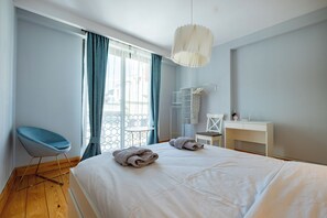 Ein Schlafzimmer mit einem großen Bett, einem Schreibtisch, einem Sessel und einem Balkon mit Vorhängen.
