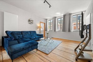 A living room with a blue sectional sofa, a glass coffee table, a lamp, and a TV on a stand. - LiT LiVING Grünstadt (Grünstadt)