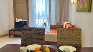 Habitación de hotel con una mesa de comedor, sillas de mimbre, un cuenco de fruta, una cama con vistas, un televisor y un espejo.