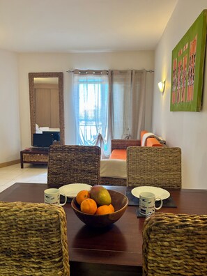 A hotel room with a dining table, wicker chairs, a bowl of fruit, a bed with a view, a television, and a mirror. - Santa Maria Studio | Relax, Pool & Kitesurf (Sal)