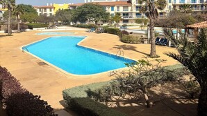 Una piscina con borde curvo, rodeada de arena y palmeras, con edificios de varios pisos al fondo.