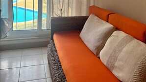 Habitación con vista a una piscina, un sofá con cojines, una lámpara de pared y una ventana con cortinas.