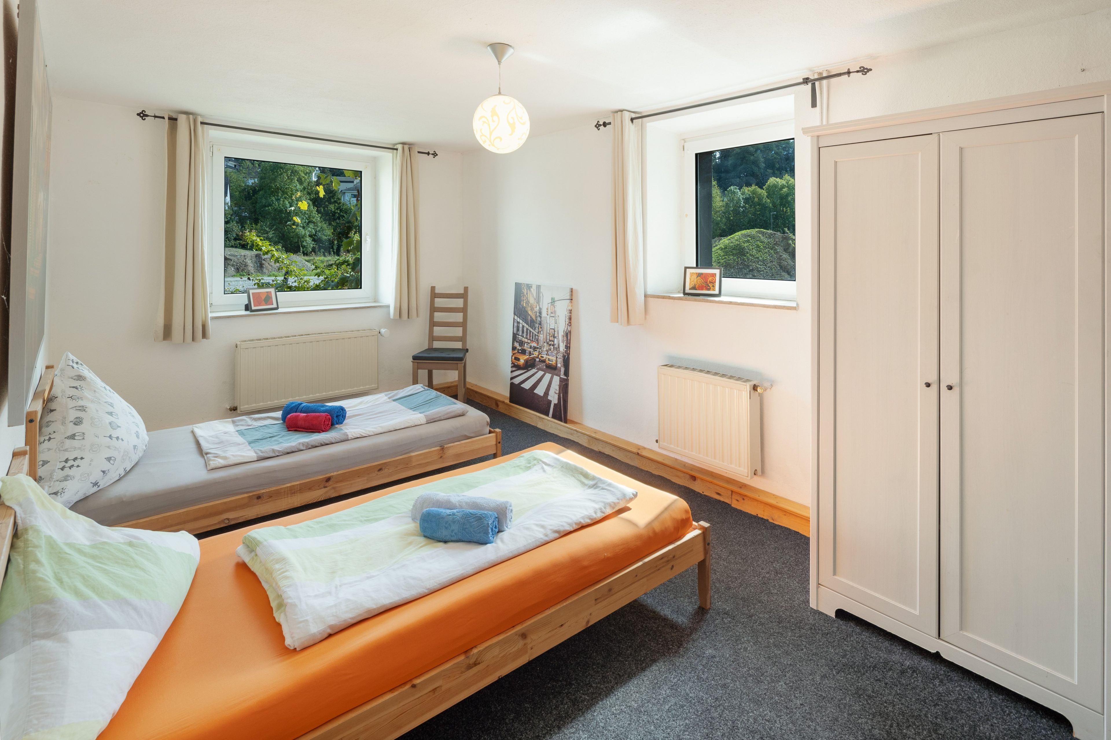 Habitación con dos camas individuales, un armario, un radiador y dos ventanas con cortinas.