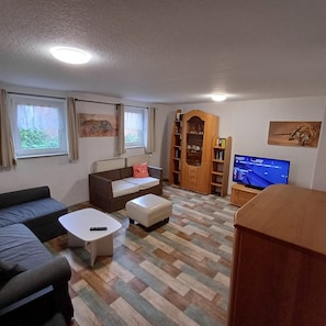 Un saló amb un sofà, una otomana, una taula de centre, televisió, una prestatgeria i una finestra amb cortines.