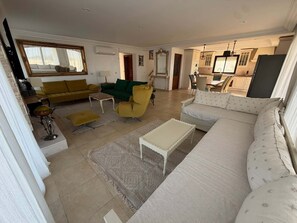 Een ruime woonkamer met een grote bank, een gele fauteuil en een groene bank. Er is een eethoek met een tafel en stoelen, en een keuken op de achtergrond.