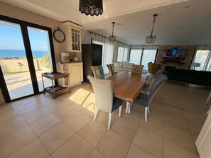 A spacious living area with a dining table, chairs, a sofa, a kitchen with a refrigerator, and a large window overlooking a scenic view.