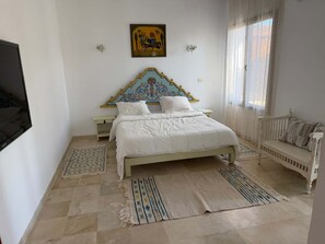 A bedroom with a bed, a nightstand, a bench, a wall-mounted TV, and a window with curtains. - Villa Bel Azur: Panoramic View of the Sea and the Golf Course (Tabarka)
