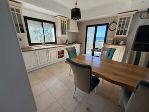 Een moderne keuken met witte kasten, een houten eettafel en uitzicht op zee door grote ramen.