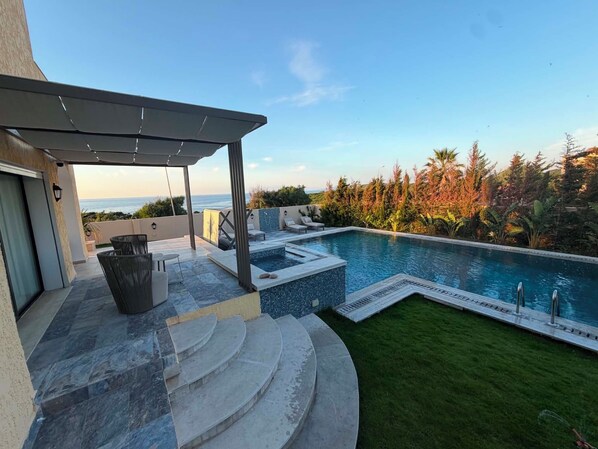 A pool with a view of the ocean, a covered patio area, and a well-maintained lawn.