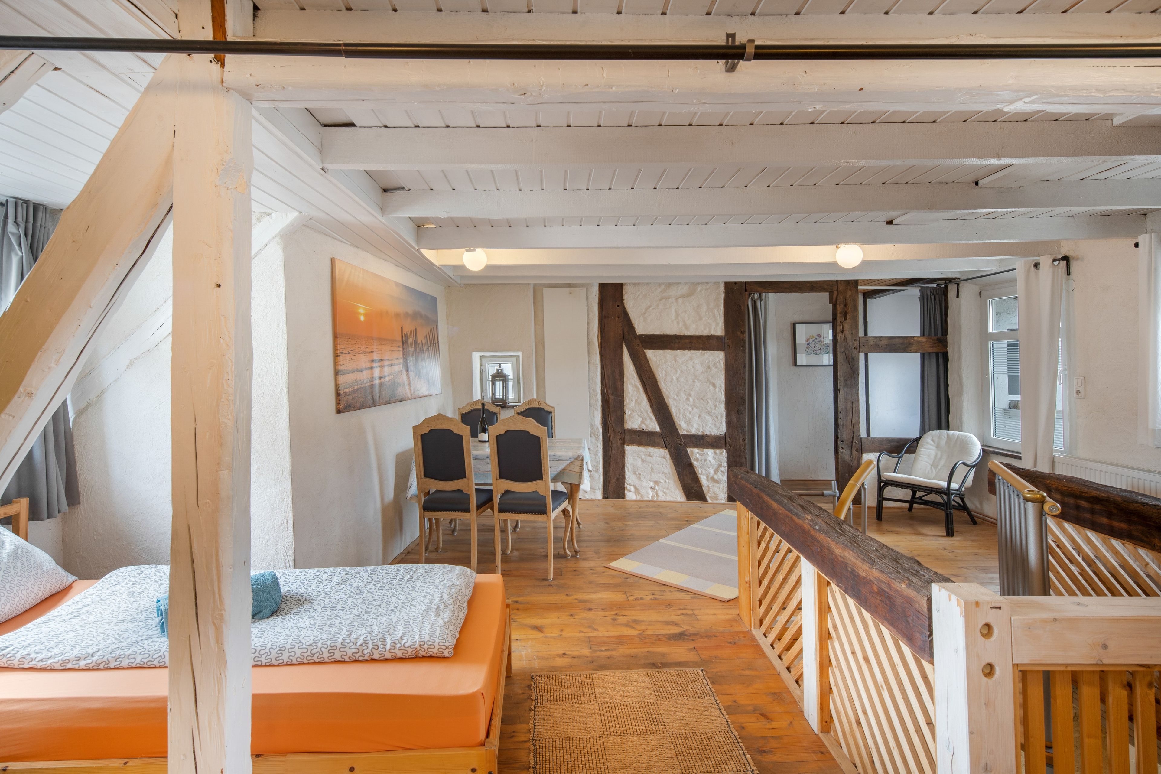 Ein Zimmer mit schrägverdeckter Decke, Holzbalken, einem Bett mit gemusterten Textilien, einem Esstisch mit Stühlen und Blick durch ein Fenster.
