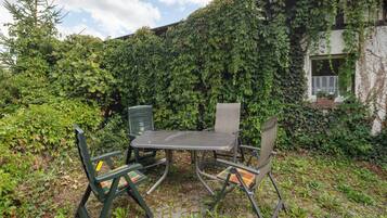 Ein Sitzbereich im Freien mit Tisch und Stühlen, umgeben von üppigem Grün und einem Gebäude mit efeuüberwachsenen Wänden.