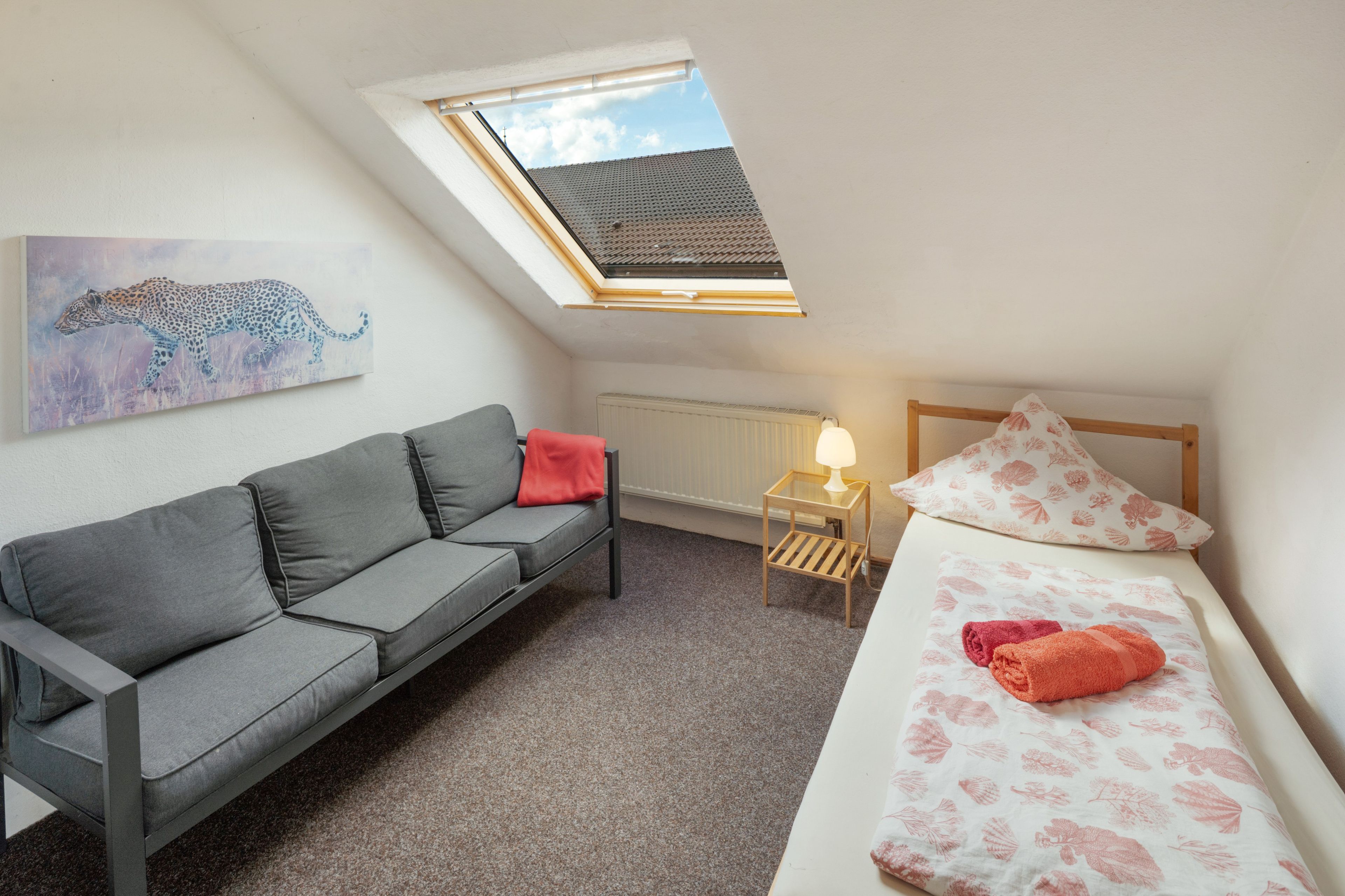 Ein kleiner, gut beleuchteter Raum mit einem grauen Sofa, einem Bett mit floralem Bettbezug, einem Oberlicht, einem Heizkörper und einem Leopardenbild an der Wand.