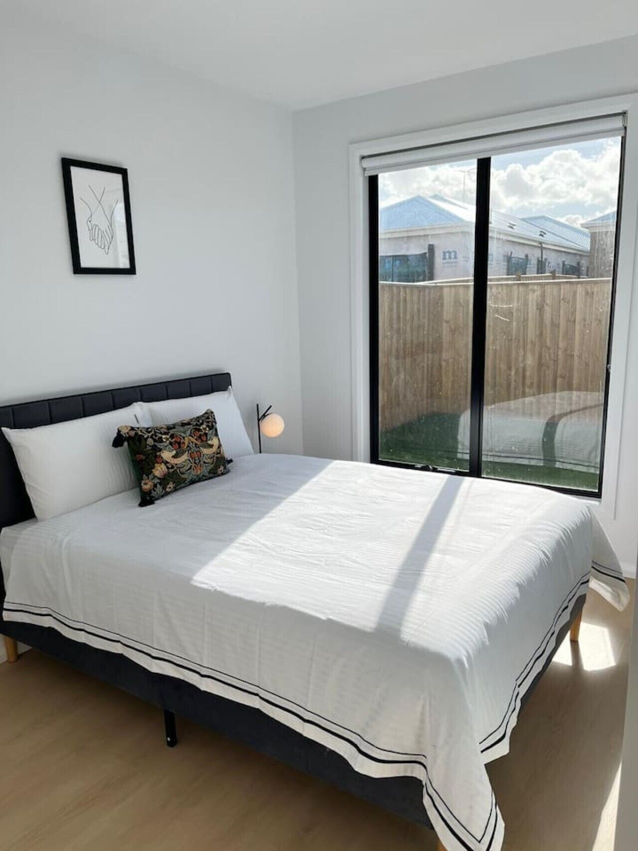 A bedroom with a bed, a bedside lamp, a framed picture, and a large window with a view of a building and a fence.