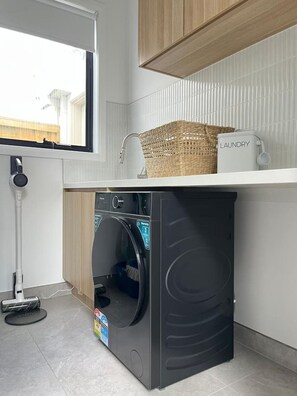 A compact laundry area with a washing machine, a drying machine, a basket, and a laundry detergent container. - Modern 4BR Family Home in Wollert, VIC (Wollert)