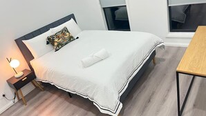 Uma cama bem arrumada com edredão branco e almofada decorativa, mesa de cabeceira com abajur, piso de madeira e vista para o exterior através de uma janela.