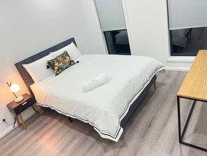 A neatly made bed with a white duvet and a decorative pillow, a bedside table with a lamp, a wooden floor, and a view of the outside through a window. - Modern 4BR Family Home in Wollert, VIC (Wollert)