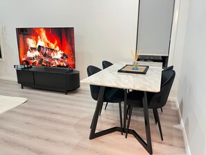 Un espace de repas moderne avec une table et des chaises, un téléviseur diffusant une image de feu de cheminée et une petite table d’appoint avec une bougie.