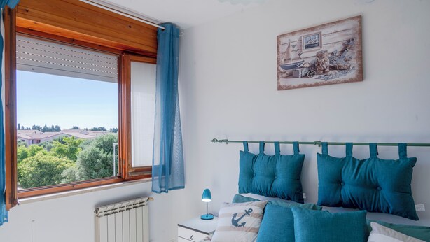 Ein Schlafzimmer mit Bett, Nachttisch, Lampe, einem Fenster mit Vorhängen und einem Wandbild.