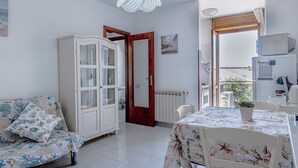 Ein Zimmer mit einer weißen Couch, einem Esstisch mit blumigem Tischläufer und einer weißen Kommode, mit Blick nach draußen.