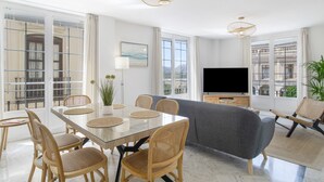 A modern living room with a glass dining table, wicker chairs, a grey sofa, and a flat-screen TV.