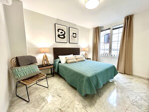 Ein Schlafzimmer mit einem Bett, zwei Nachttischlampen, einem Sessel mit Kissen, einem Fenster mit Vorhängen und zwei gerahmten Bildern an der Wand.