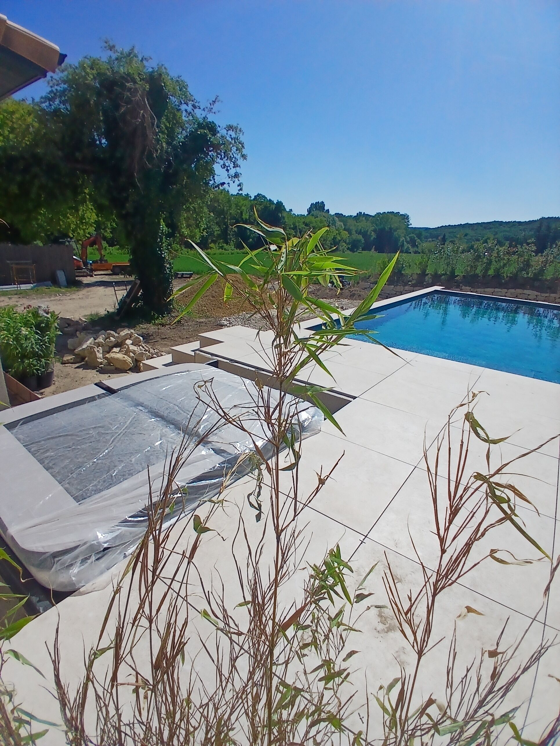 Une piscine, avec un ciel bleu clair en arrière-plan, entourée de verdure et bordée d’un chantier de construction.