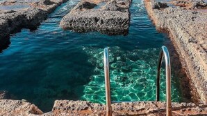 Una costa rocciosa con acque limpide, una scala di metallo e velieri all'orizzonte.