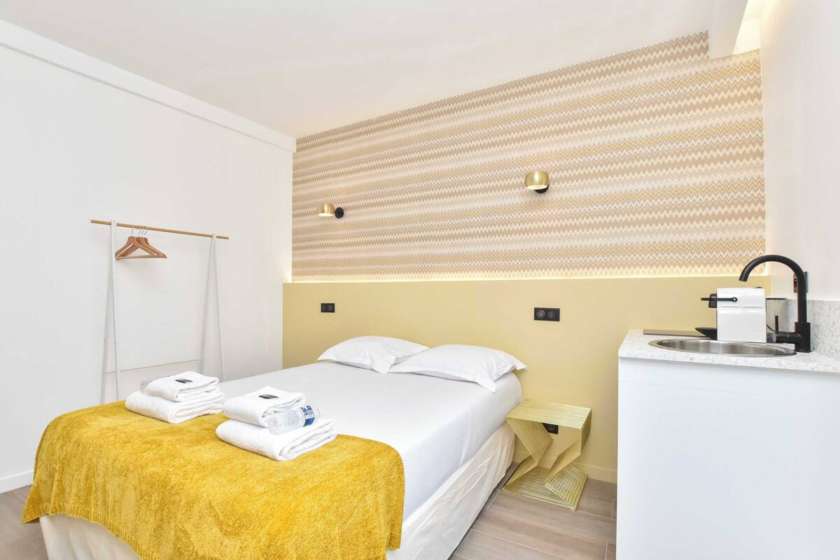 A hotel room with a bed, a bedside table, a towel rack, and a wall-mounted light fixture.