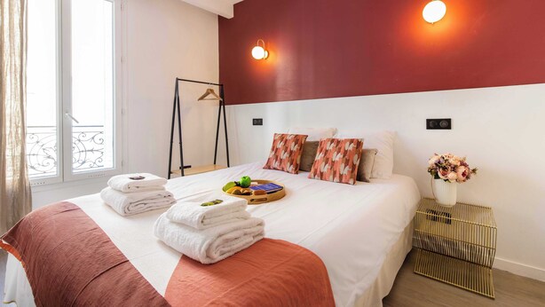 A neatly made bed with towels, a tray with fruits and a book, a hanging coat rack, and a vase with flowers on a bedside table. - Palette Paris Tour Eiffel (Paris)