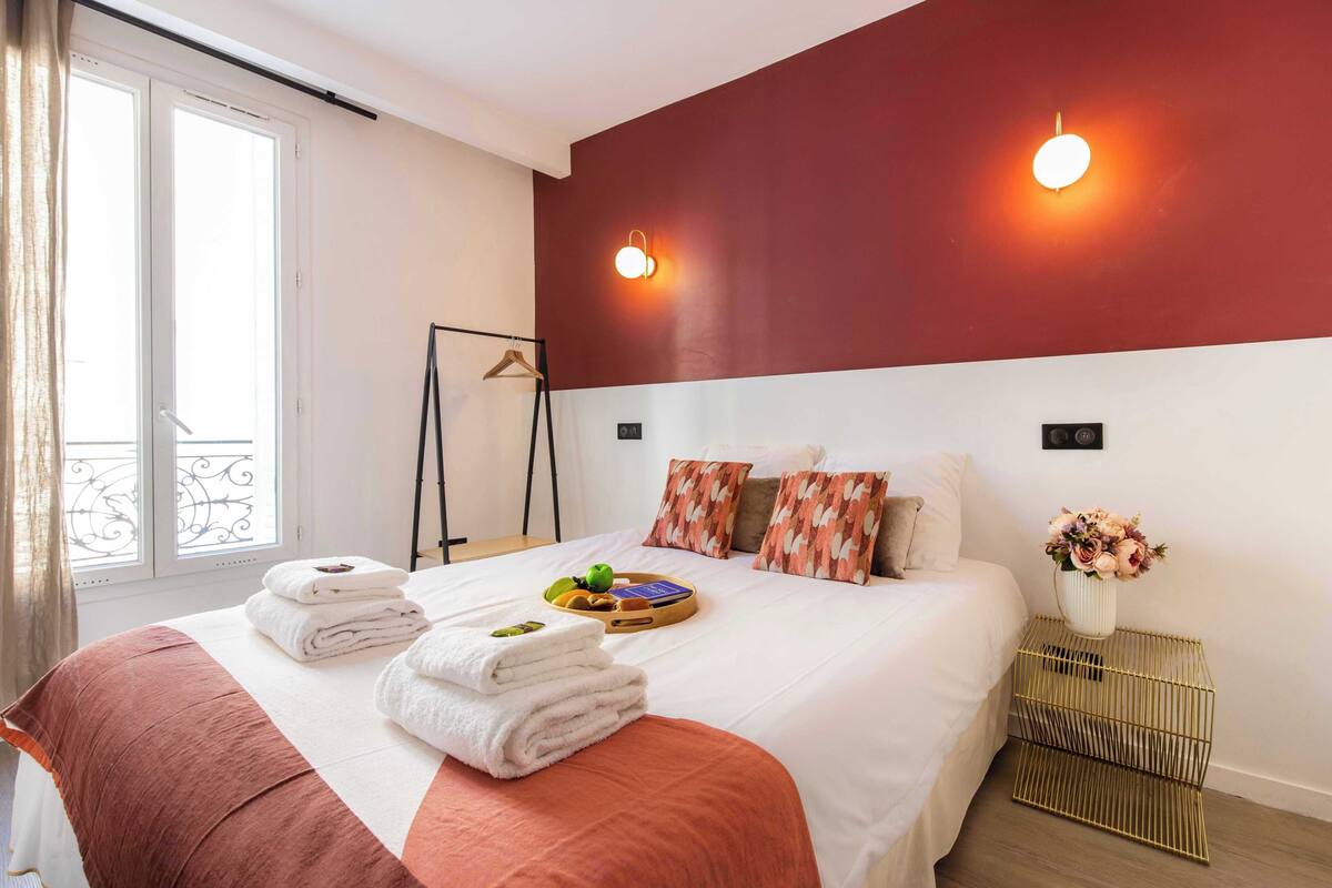 A neatly made bed with towels, a tray with fruits and a book, a hanging coat rack, and a vase with flowers on a bedside table.