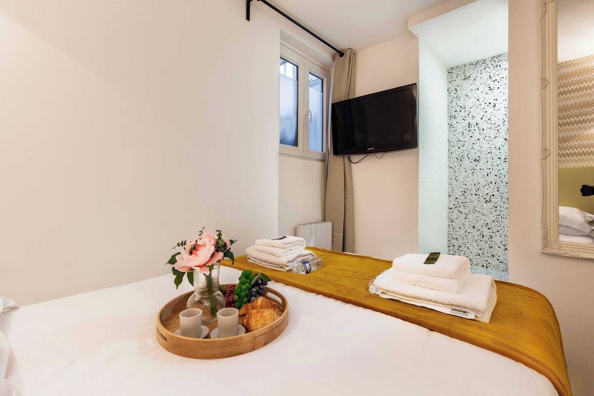 A hotel room with a bed, a wooden side table with towels and a tray of breakfast items, a TV mounted on the wall, and a window with curtains.