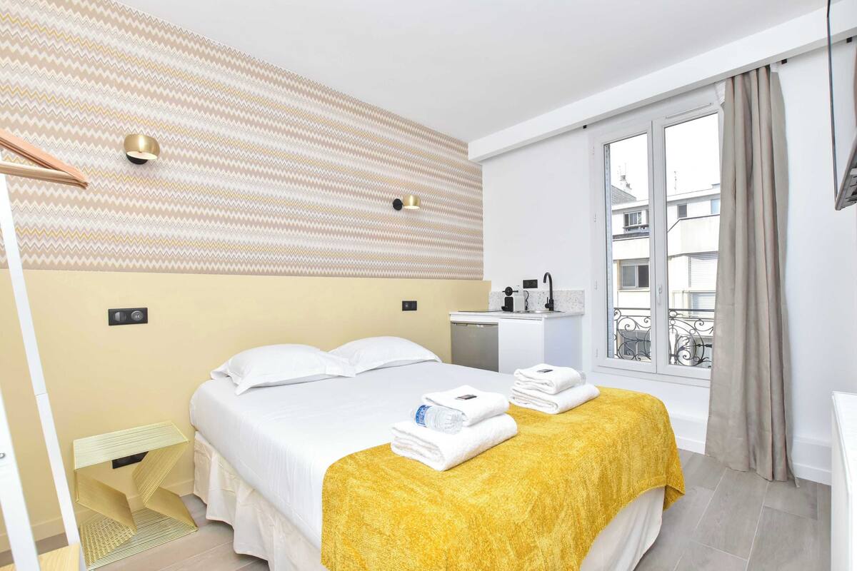 A hotel room with a bed, a bedside table, a window with curtains, and a view of buildings outside.