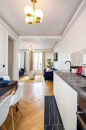 Eine moderne Küche mit Marmorarbeitsplatte, ein Essbereich mit weißem Tisch und Stühlen sowie ein Wohnzimmer mit Sofa und Pflanze.