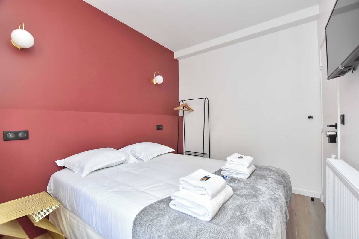 A hotel room with a bed, white linens, a grey blanket, a wooden side table, a mirror, and a TV mounted on the wall.