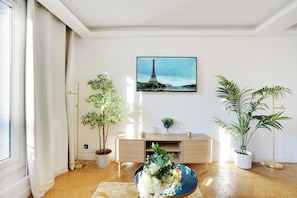 A living room with a large window, a wooden floor, a white sofa, a coffee table with flowers, and a painting of the Eiffel Tower.