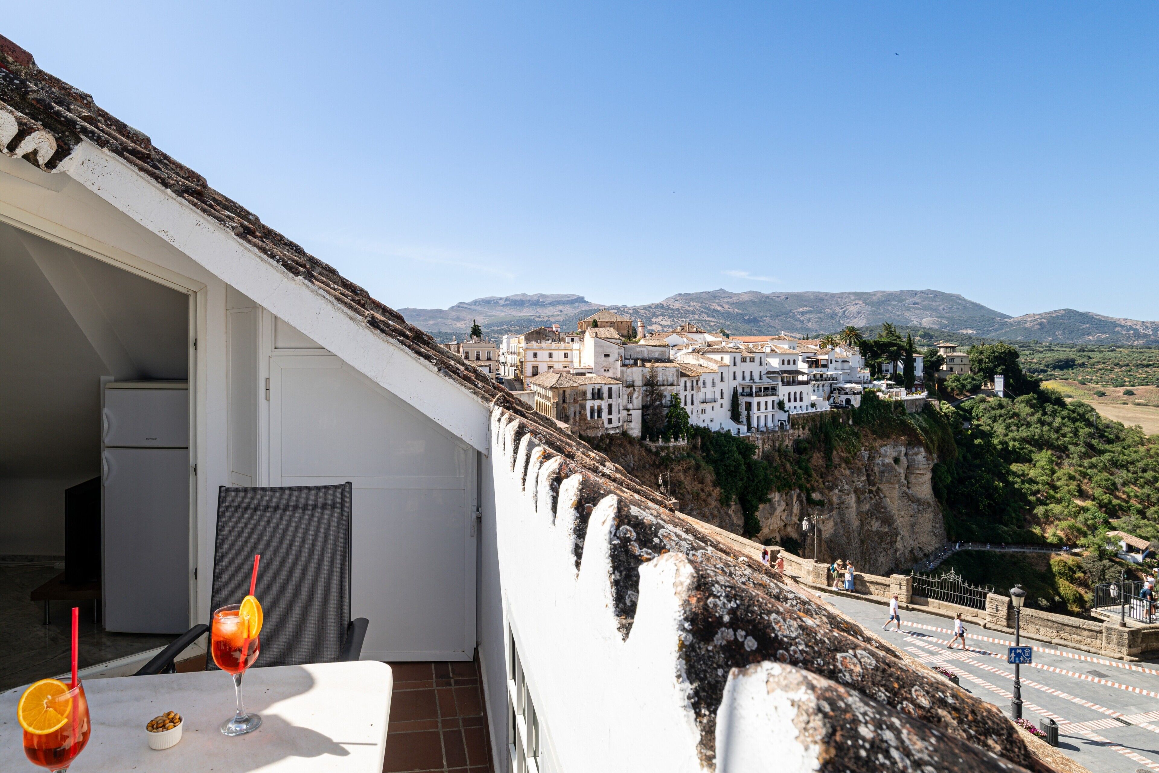 A balcony with a view of a town, a table with drinks, and a refrigerator.