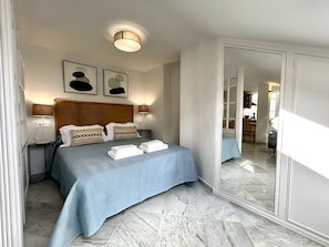 Habitación de hotel con cama, lámparas de noche, cabecero, espejo y vista a la habitación contigua.