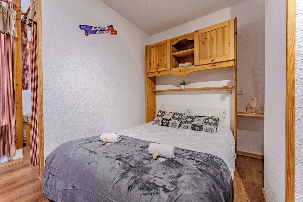 A bedroom with a bed, a wooden shelf above the bed, and a checkered curtain on the left.