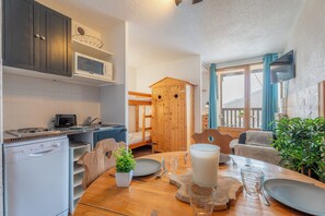 A compact kitchen with a microwave, stove, and dishwasher. There is a dining table with a candle, plates, and glasses. A wooden cabinet and a plant are also visible. - Cimes de Caron Val Thorens (Val Thorens)
