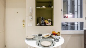 Un coin repas avec une table dressée pour deux personnes, une petite étagère verte où sont rangés des appareils de cuisine, et une fenêtre donnant sur un beau paysage.