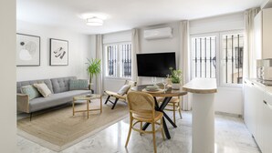 Ein modernes Wohnzimmer mit Sofa, Couchtisch, Esstisch und Stühlen. An der Wand ist ein Flachbildfernseher angebracht, eine Klimaanlage und ein Fenster mit Blick.